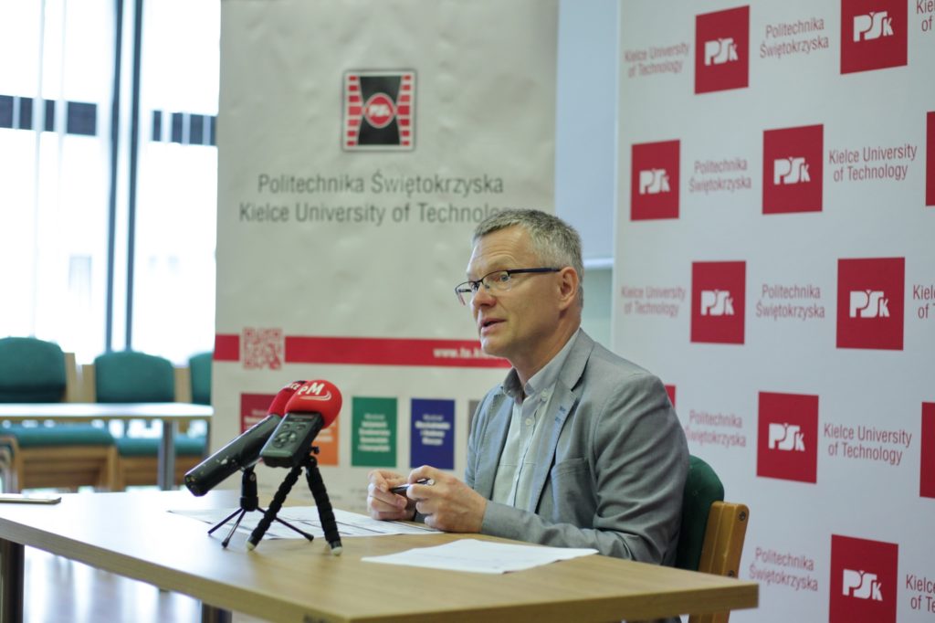 Mężczyzna w okularach siedzi przy stole z mikrofonami. W tle logo Politechniki Świętokrzyskiej.