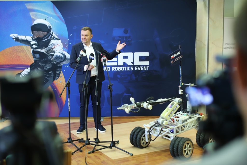 Mężczyzna w garniturze przemawia przy mikrofonach obok robota. W tle grafika astronauty i logo 