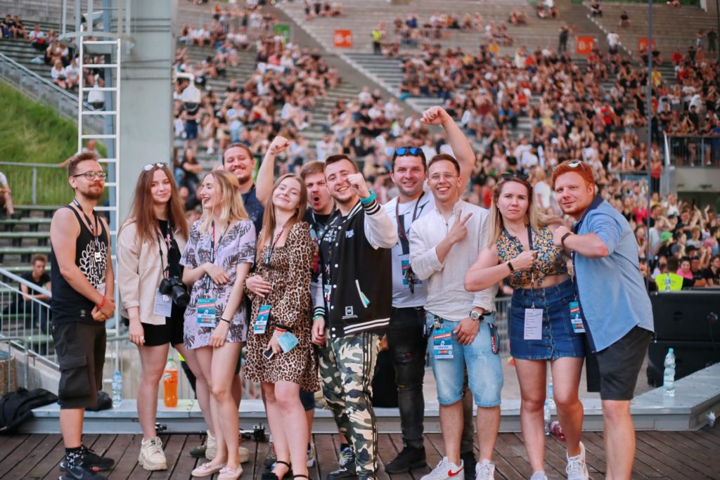 Grupa młodych ludzi pozuje do zdjęcia na tle tłumu na widowni. Wszyscy są uśmiechnięci i radośni.
