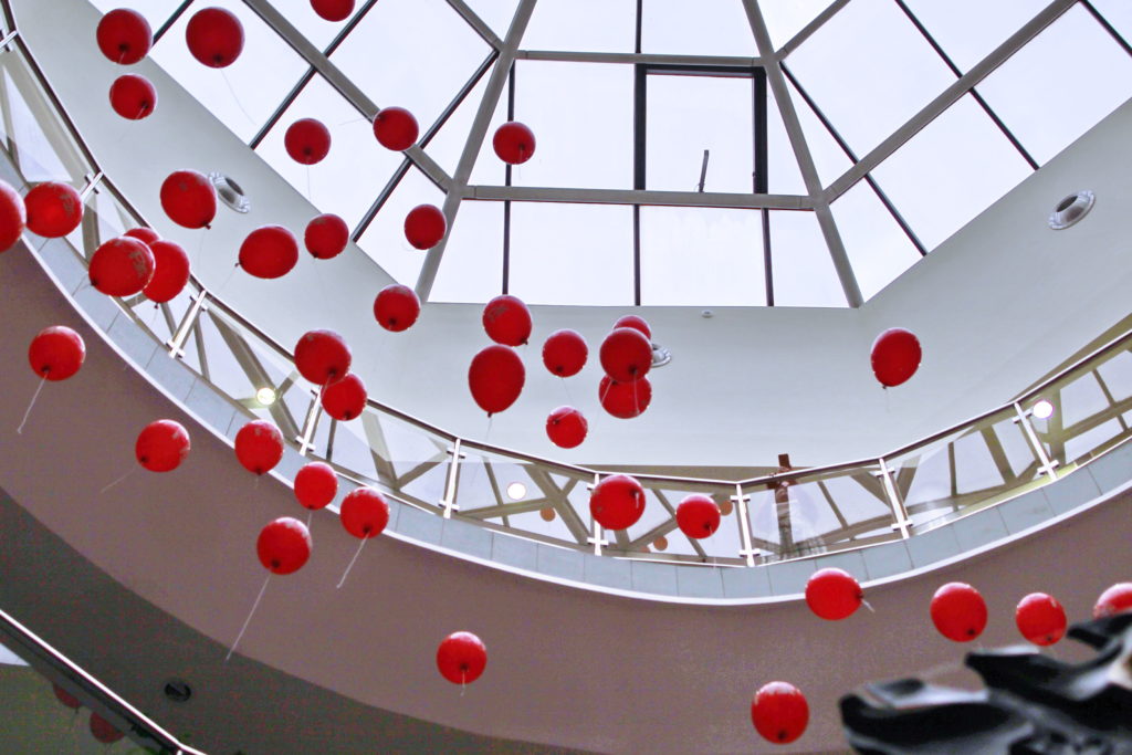 Czerwone balony unoszą się pod przeszklonym sufitem centrum handlowego. Widoczne balony, balustrady, okna.
