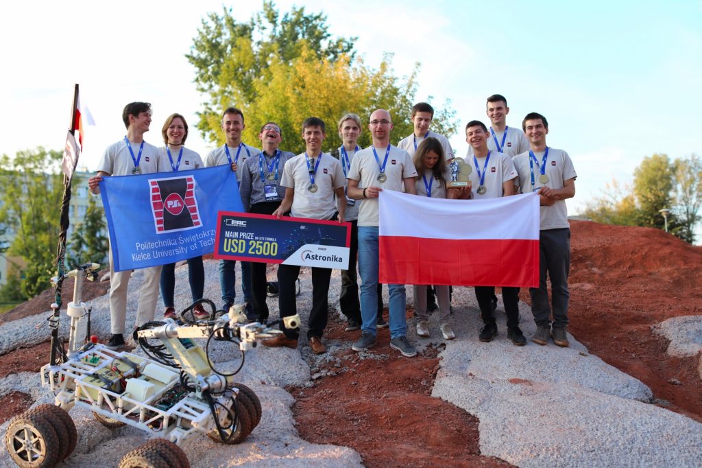 Grupa młodych ludzi z Politechniki Świętokrzyskiej świętuje wygraną z robotem. Kluczowe obiekty: robot, flagi, nagrody.