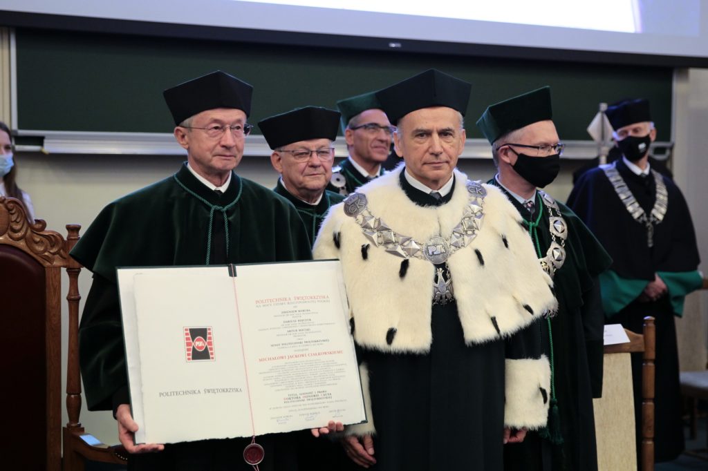 Uroczystość wręczenia dyplomu. W centrum mężczyzna w futrzanym płaszczu i łańcuchu. Kluczowe obiekty: dyplom, togę, łańcuch.