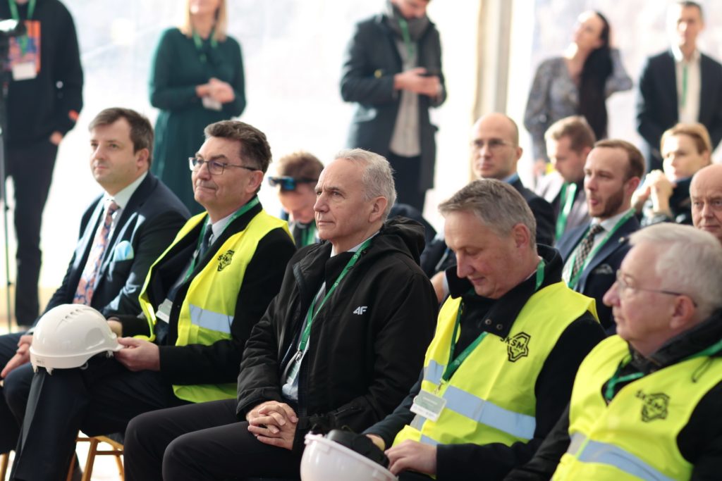 Mężczyźni w kamizelkach odblaskowych i garniturach siedzą. W tle ludzie. Kluczowe obiekty: kamizelki, garnitury, kaski.