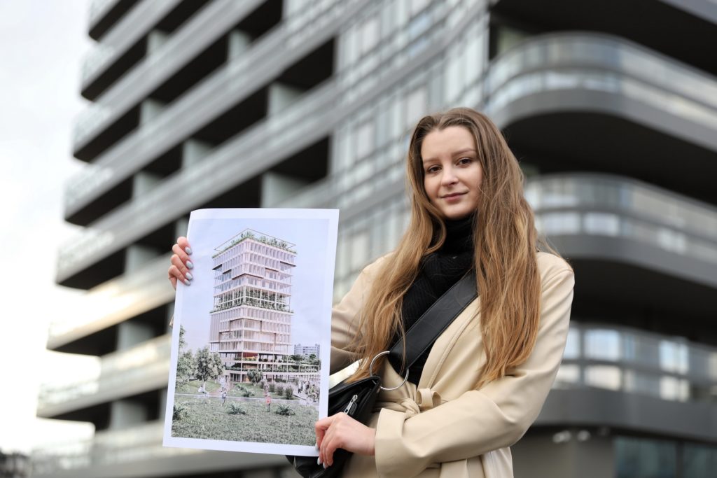 Kobieta trzyma wizualizację nowoczesnego budynku. W tle widać architekturę miejską.