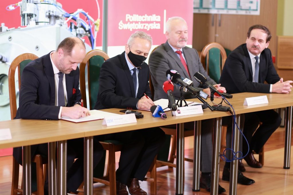 Czterech mężczyzn w garniturach siedzi przy stole, podpisując dokumenty.