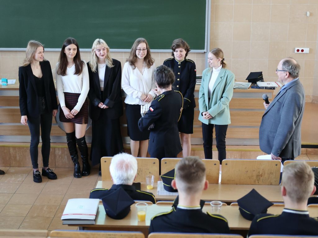 Grupa osób stoi przed tablicą, jeden mężczyzna przemawia. Wśród nich są młode kobiety w strojach galowych i garniturach.