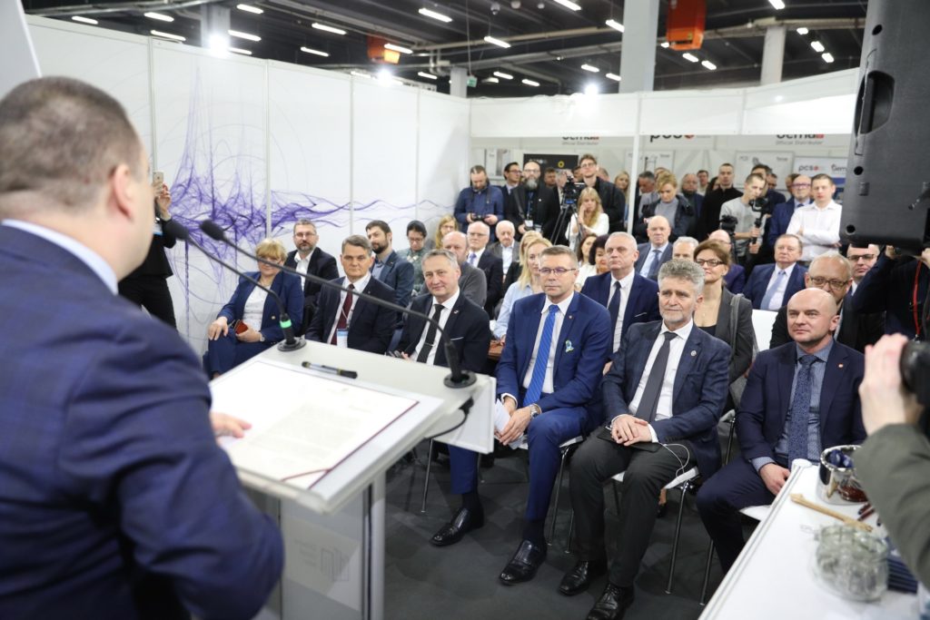 Mężczyzna w garniturze przemawia do zgromadzonych osób. W tle widać uczestników wydarzenia.