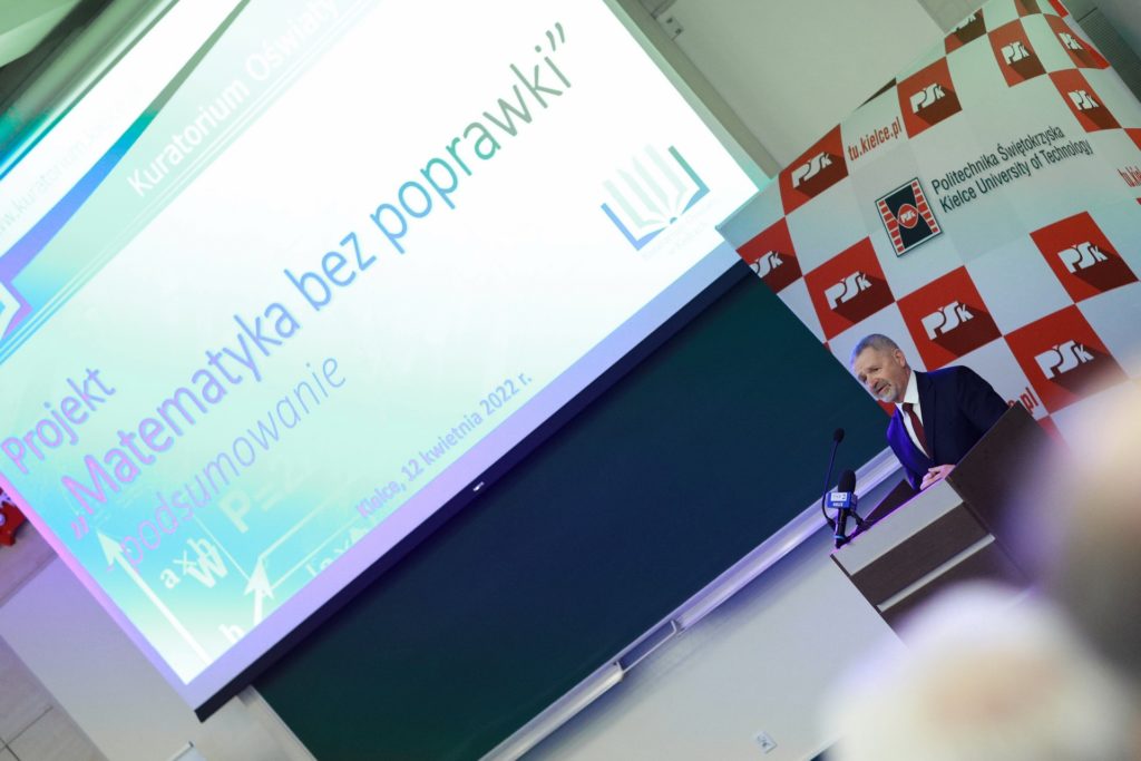 Mężczyzna w garniturze przemawia na tle ekranu z napisem 