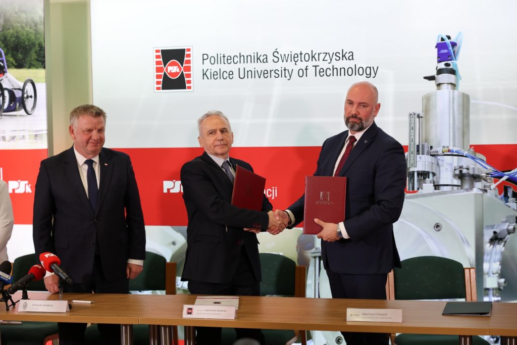 Trzech mężczyzn w garniturach ściska sobie dłonie, trzymając dokumenty. W tle logo Politechniki Świętokrzyskiej.