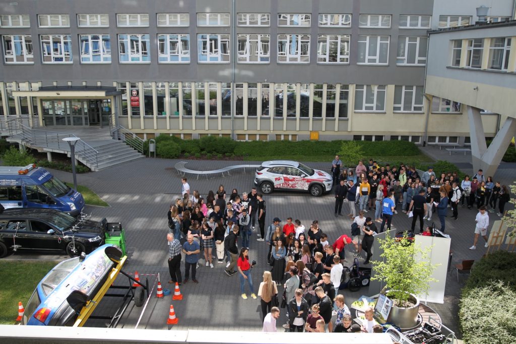 Grupa ludzi stoi przed budynkiem, obok samochodów i stoisk. Kluczowe obiekty: ludzie, samochody, budynek, stoiska.