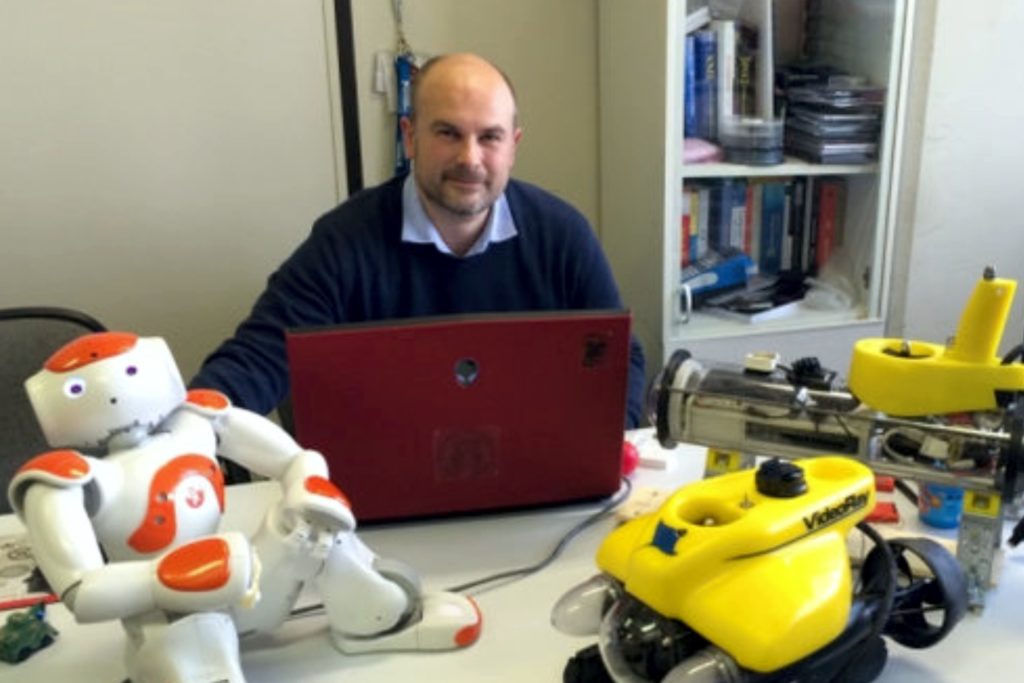 Mężczyzna z robotami i laptopem. Na stole robot humanoidalny, żółty pojazd podwodny i czerwony laptop.