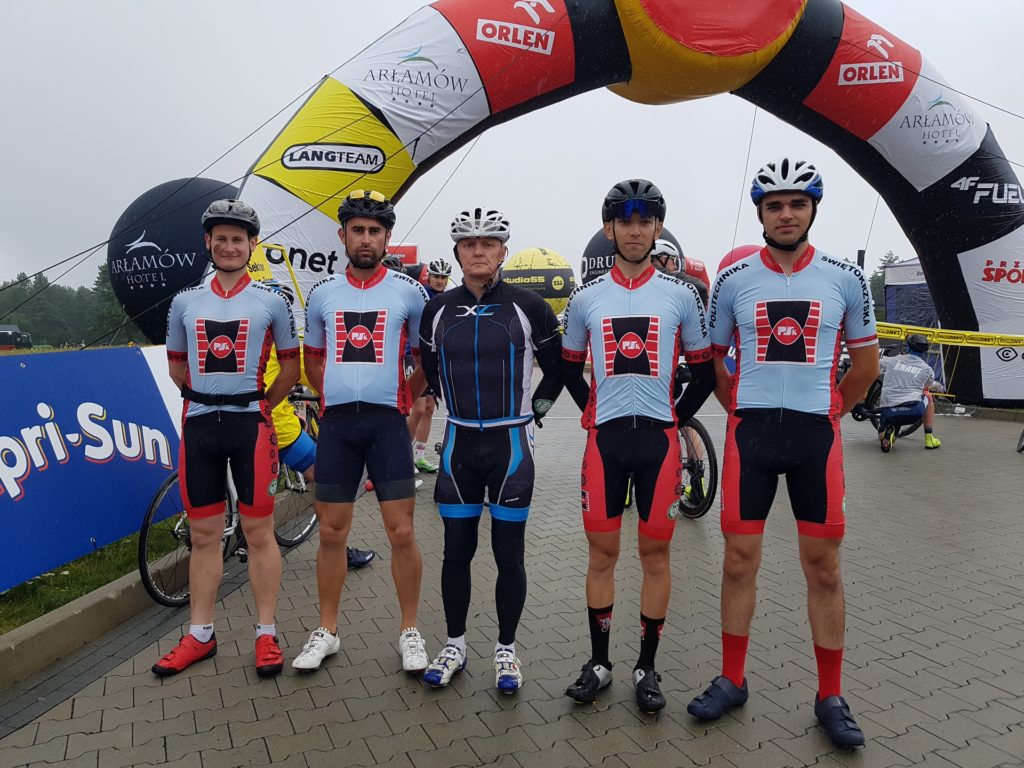 Kolarze w strojach sportowych stoją pod bramą startową. Kluczowe obiekty: kolarze, kaski, stroje, brama startowa, sponsorzy.