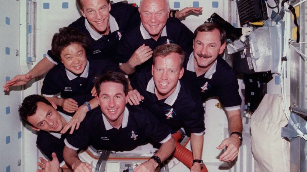 Siedmiu astronautów w ciemnych koszulkach z logo STS-95 Discovery. Kontekst: misja kosmiczna.