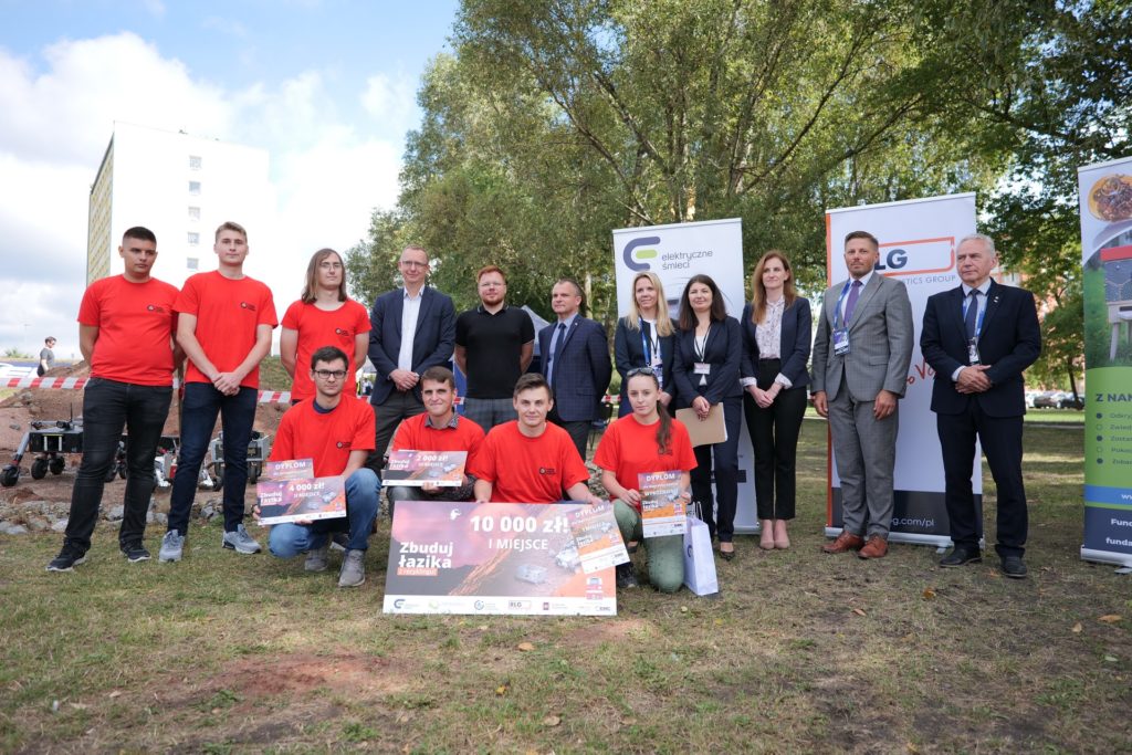 Grupa osób w czerwonych koszulkach i garniturach pozuje z dyplomami i nagrodami pieniężnymi.