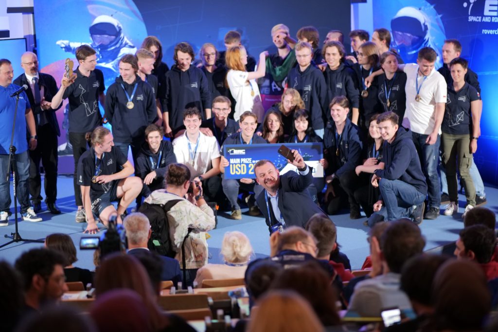 Grupa młodych ludzi w strojach z logo, odbiera nagrodę. W tle ekran z astronautą.