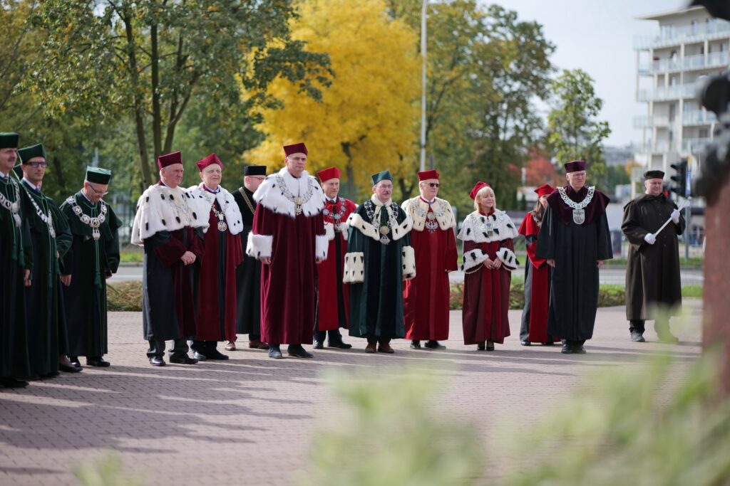 Grupa osób w togach akademickich stoi na zewnątrz. Kluczowe obiekty: togi, czapki, łańcuchy. Kontekst: uroczystość akademicka.