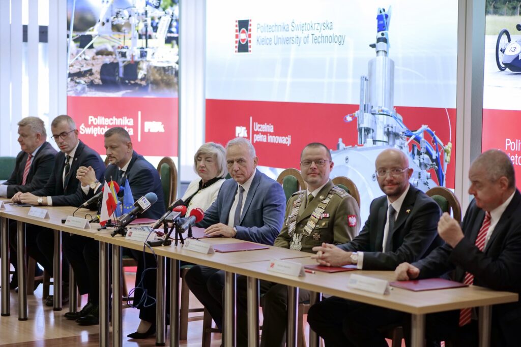 Ludzie siedzą przy stole konferencyjnym z flagami i mikrofonami. Kontekst: spotkanie w Politechnice Świętokrzyskiej.