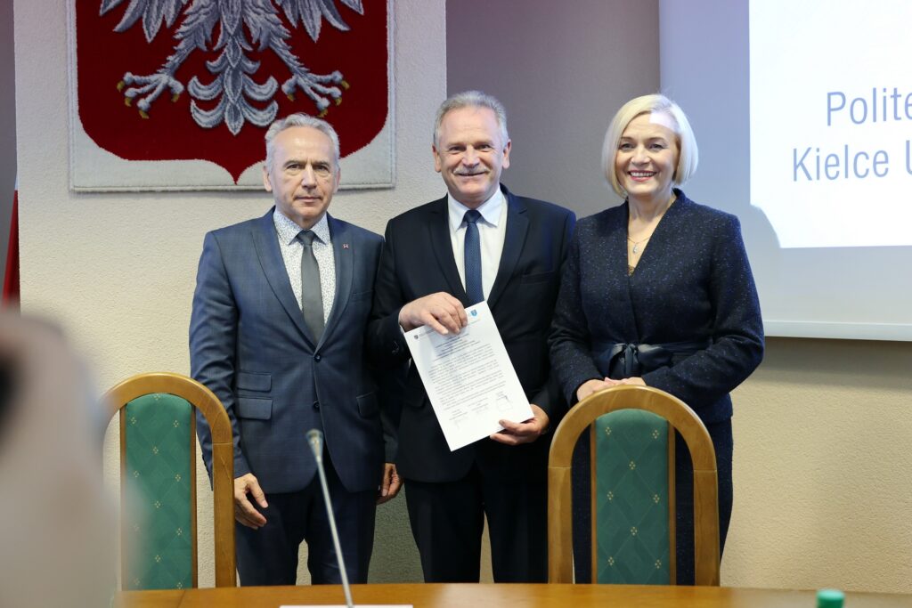 Trzy osoby w eleganckich strojach stoją przed herbem Polski. Mężczyzna w środku trzyma dokument.