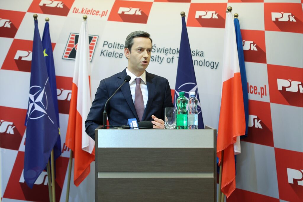 Mężczyzna w garniturze przemawia za mównicą. W tle flagi NATO, Polski i UE. Kontekst: konferencja prasowa.