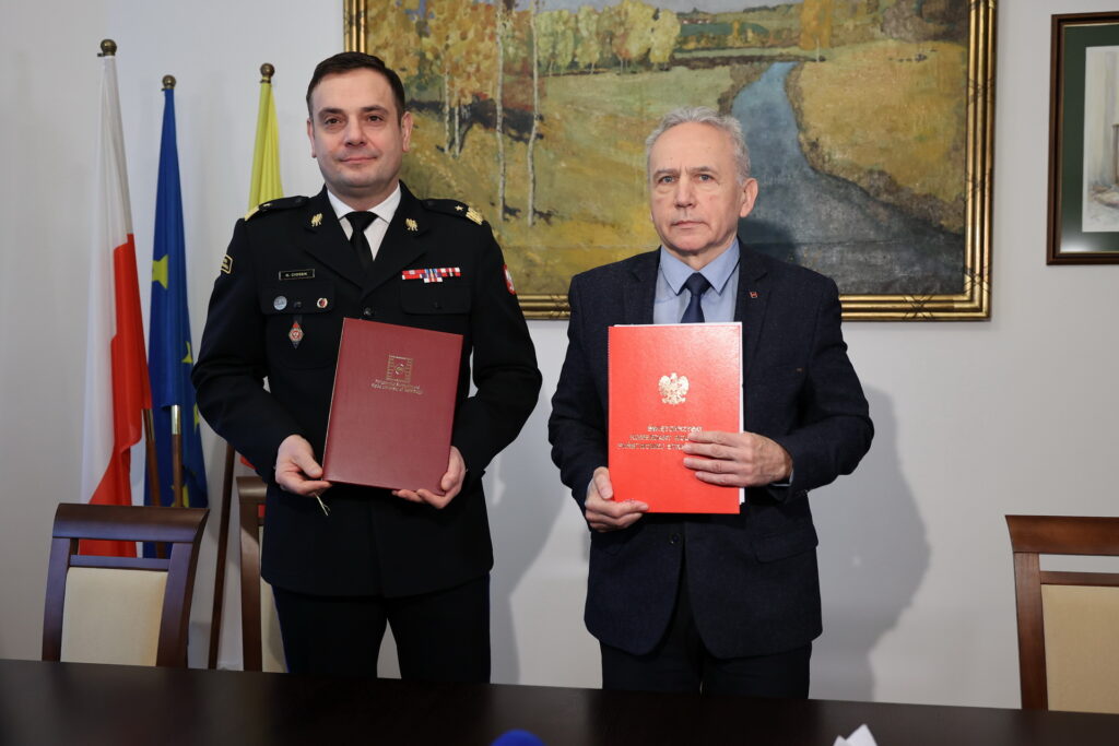 Dwóch mężczyzn trzyma dokumenty. Jeden w mundurze, drugi w garniturze. W tle flagi i obraz.