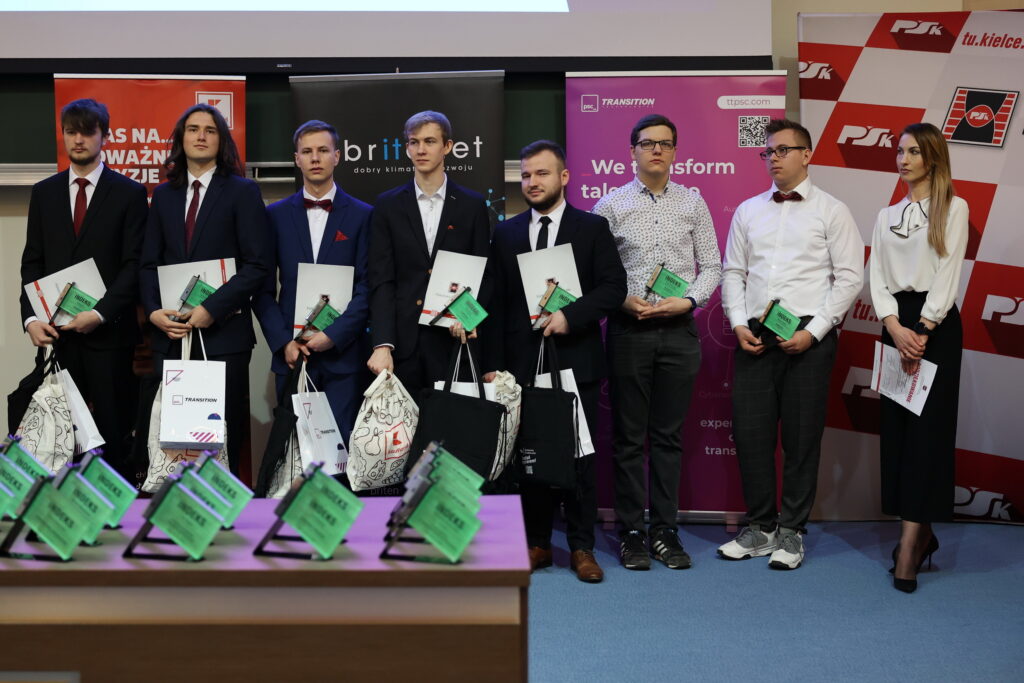 Osoby w garniturach i sukience stoją na scenie, trzymając dyplomy i torby. W tle banery.