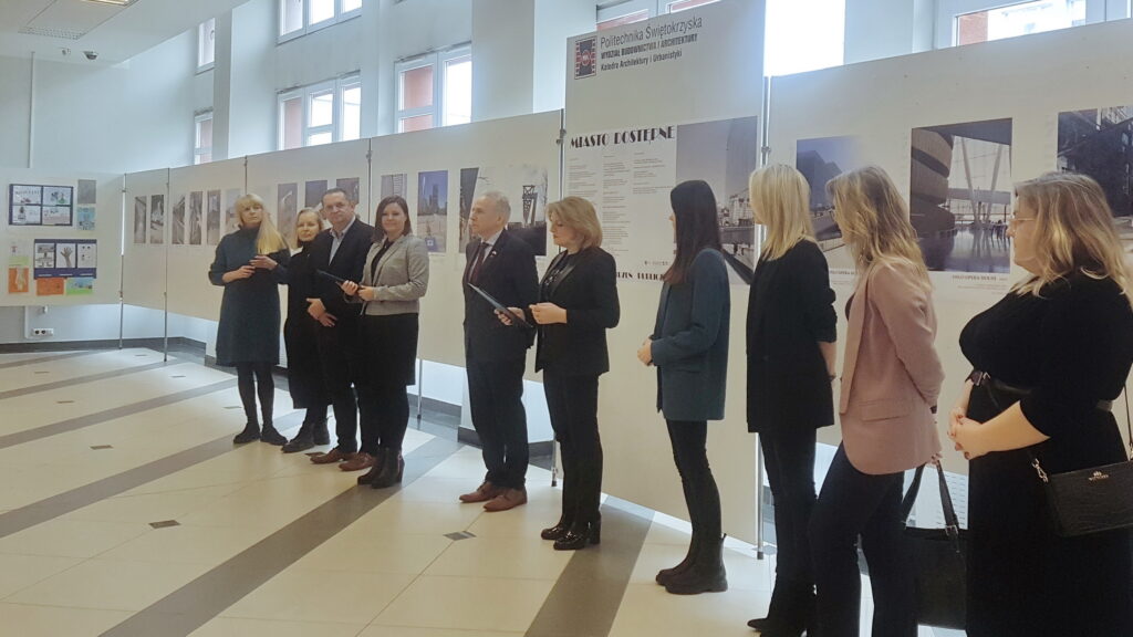 Grupa osób stoi przed wystawą prac architektonicznych. Wystawa zawiera plakaty z podpisami, m.in. 