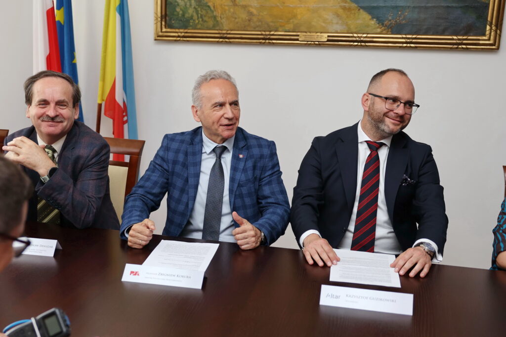 Trzech mężczyzn w garniturach siedzi przy stole. Flagi i obraz wiszą na ścianie. Kontekst: spotkanie.