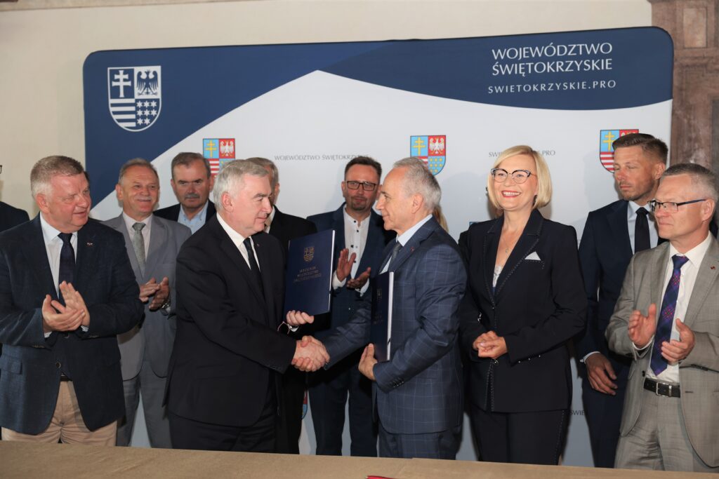 Mężczyźni i kobieta w garniturach stoją przed banerem z herbem.