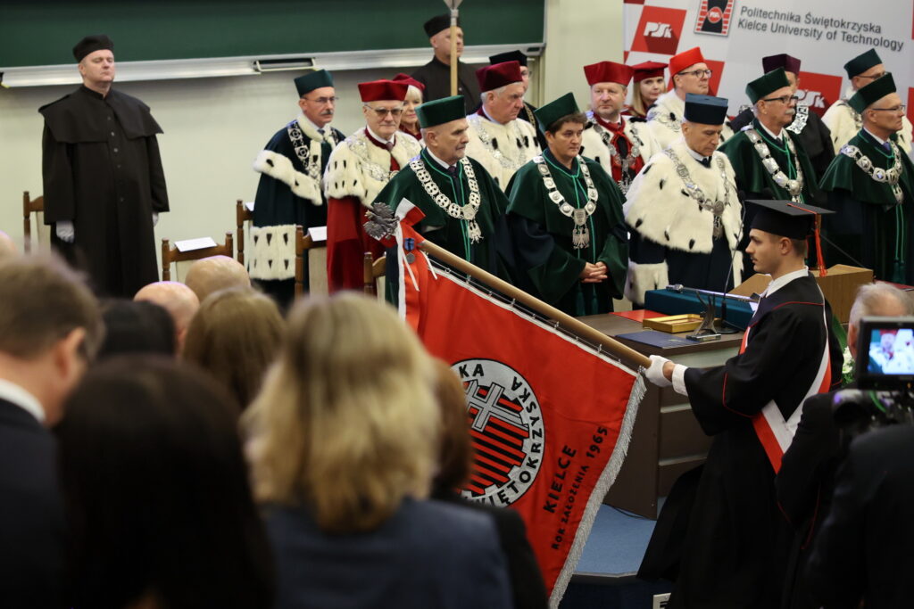 Uroczystość akademicka z udziałem osób w togach. Widoczny sztandar z napisem 