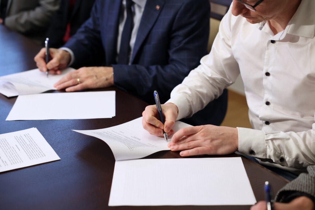 Ludzie podpisują dokumenty na ciemnym stole. Kluczowe obiekty: dokumenty, długopisy, dłonie.