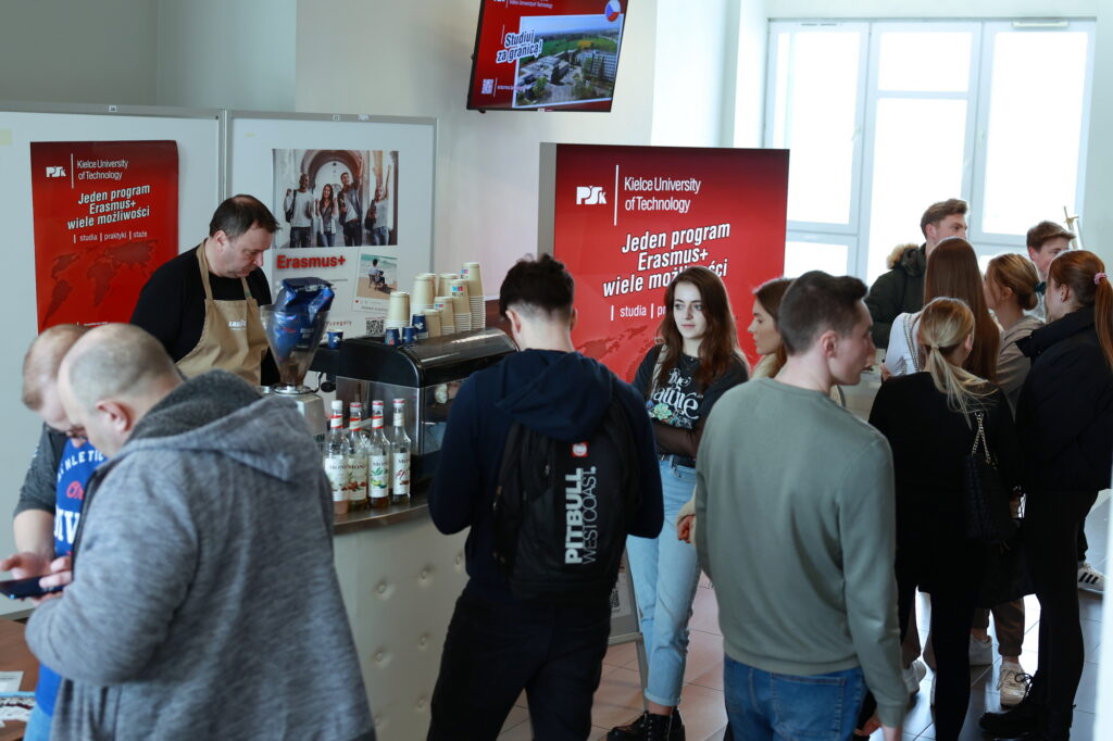Ludzie stoją przy stoisku z kawą, na tle banerów promujących program Erasmus+.
