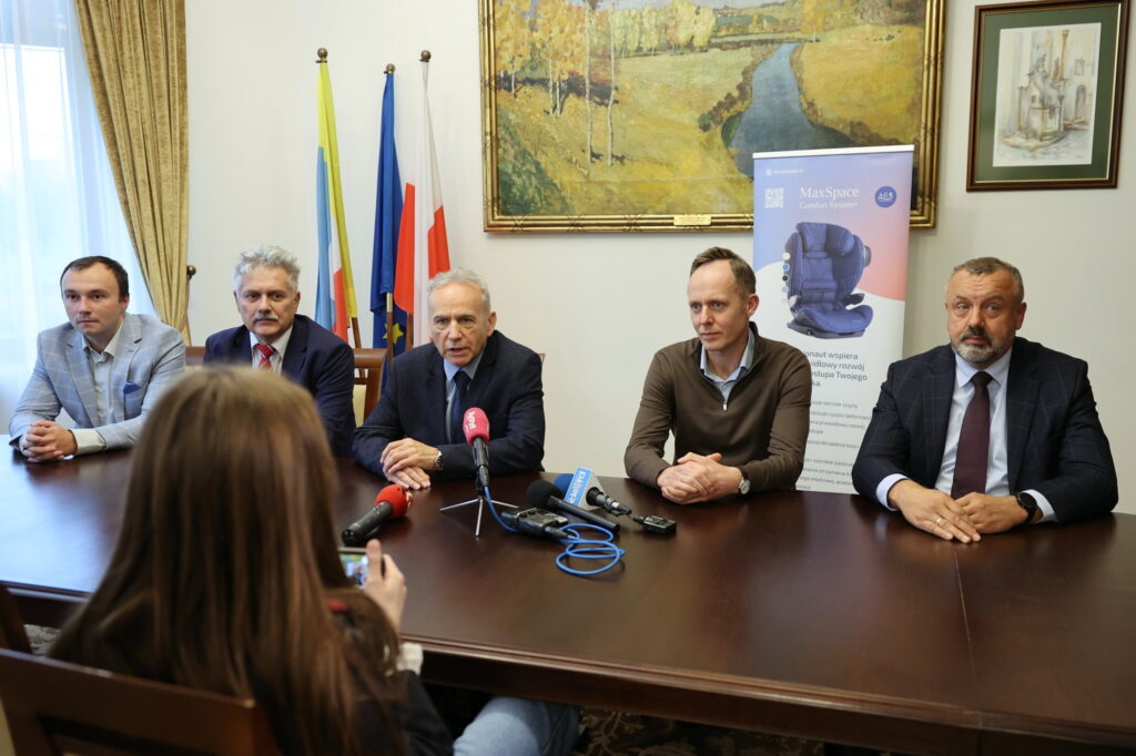 Pięciu mężczyzn siedzi przy stole, przed nimi mikrofony i baner. Kontekst: konferencja prasowa.