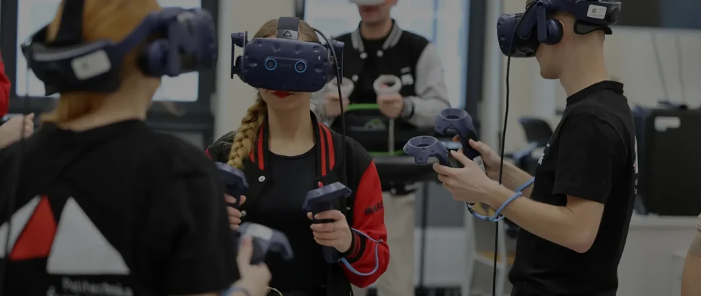 Osoby w goglach VR i kontrolerach w rękach, w pomieszczeniu z wyposażeniem. Kontekst: technologia, edukacja.