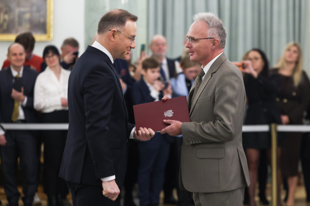 Dwóch mężczyzn w garniturach wręcza sobie dokument. W tle widać ludzi.