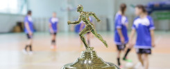 Złoty puchar z postacią biegacza na tle hali sportowej z zawodniczkami w niebieskich strojach.