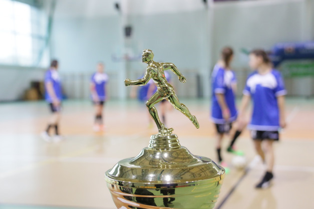 Złoty puchar z postacią biegacza na tle hali sportowej z zawodniczkami w niebieskich strojach.