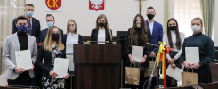 Grupa osób w maseczkach stoi w sali z herbami i krzyżem. Kluczowe obiekty: ludzie, dokumenty, hulajnoga, torby.