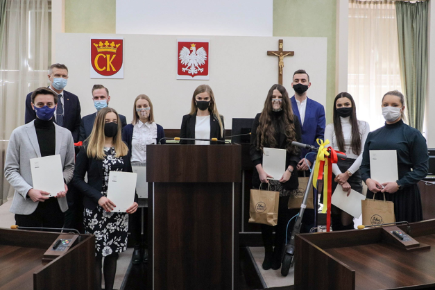 Grupa osób w maseczkach stoi w sali z herbami i krzyżem. Kluczowe obiekty: ludzie, dokumenty, hulajnoga, torby.