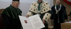 Trzech mężczyzn w togach akademickich. Dwóch z nich trzyma dokument. Kontekst: uroczystość akademicka.
