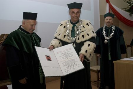 Trzech mężczyzn w togach akademickich. Dwóch z nich trzyma dokument. Kontekst: uroczystość akademicka.
