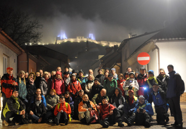 Grupa ludzi pozuje do zdjęcia nocą przed zamkiem. Widać zamek, budynki i znak drogowy.