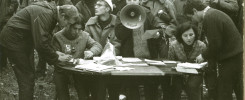 Grupa ludzi przy stole z dokumentami. Kobieta z megafonem, mężczyzna w czapce, kobiety i mężczyźni w ubraniach z epoki.