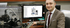 Mężczyzna w garniturze siedzi przy biurku z komputerem wyświetlającym projekt techniczny.