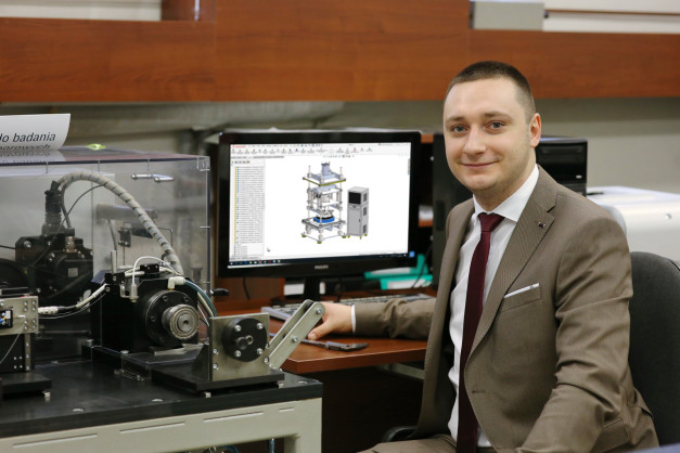 Mężczyzna w garniturze siedzi przy biurku z komputerem wyświetlającym projekt techniczny.