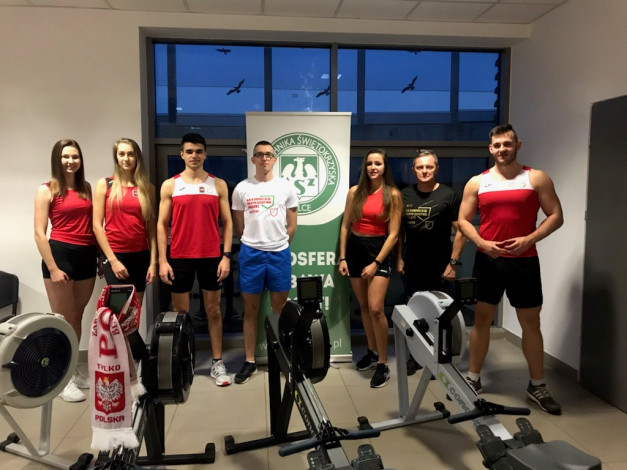 Grupa osób w strojach sportowych stoi przed maszynami wioślarskimi. W tle baner z logo.