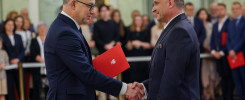 Dwóch mężczyzn w garniturach ściska sobie dłonie. Jeden z nich trzyma czerwoną teczkę z godłem.