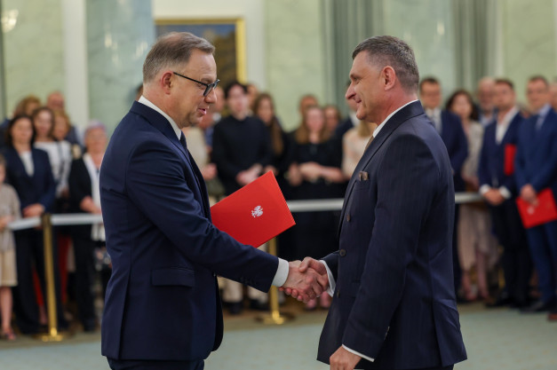 Dwóch mężczyzn w garniturach ściska sobie dłonie. Jeden z nich trzyma czerwoną teczkę z godłem.