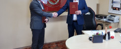 Dwóch mężczyzn w garniturach ściska sobie dłonie, trzymając dokumenty. W tle widać logo szkoły i kalendarz.