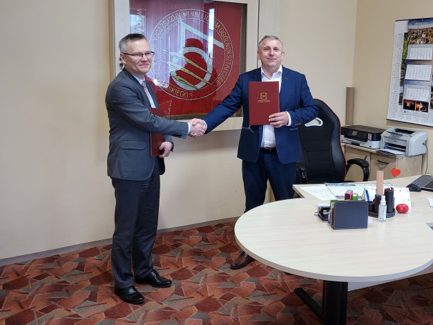 Dwóch mężczyzn w garniturach ściska sobie dłonie, trzymając dokumenty. W tle widać logo szkoły i kalendarz.