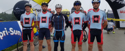 Kolarze w strojach sportowych stoją pod bramą startową. Kluczowe obiekty: kolarze, kaski, stroje, brama startowa, sponsorzy.